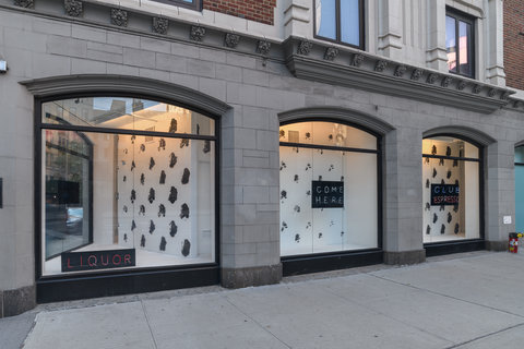 The exterior of a light colored building at street level with three large windows. In the center of the windows are small black signs, behind the signs is a black and white spotted background.