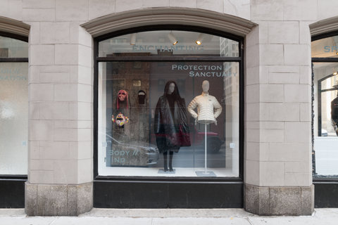 Image of two mannequins in the exhibition. On the right side of the image, an ornate fabric is draped and three mannequin heads with hats and masks are mounted on it. In the middle, a mannequin wears a very puffy black coat and black tights. On the right, a mannequin wears a light, white, puffer jacket with its hands on its hips. The mannequin on the right does not have legs.