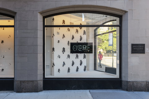 A center view of a window on street level. Inside the window display is a black and white spotted background with a small black sign. In the center the sign reads the words: come here. The letters overlap each other, in blue neon lights.