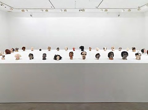 A large collection of small sculpted heads, all facing outward, are displayed on a massive white platform.