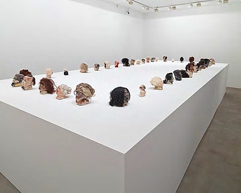 A large collection of small sculpted heads, all facing outward, are displayed on a massive white platform.