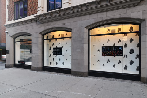 The exterior of a light colored building at street level with three large windows. In the center of the windows are small black signs, behind the signs is a black and white spotted background.