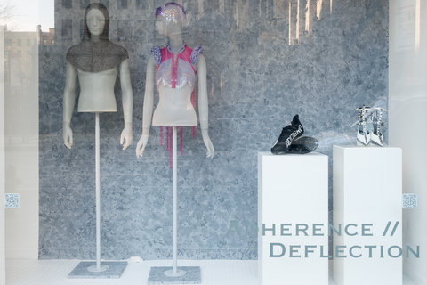 Image of two mannequins in the exhibition and two pairs of shoes. On the left, a mannequin wears a grey hood, and nothing else. The mannequin is missing the lower half of the body. Next to this mannequin, another mannequin wears a pink and purple crocheted item around their head and shoulders. In the foreground, through the window, a pair of black sneakers sits on a pedestal next to a pair of white heels with silver spikes along the straps. 