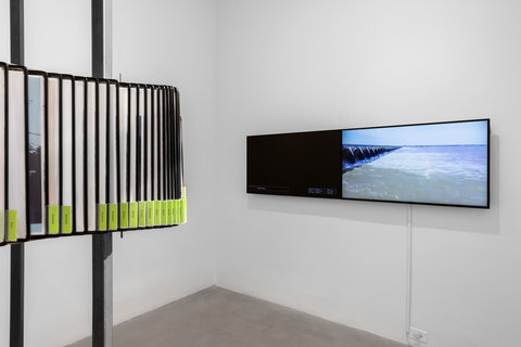 Installation view of the exhibition featuring flippable archival papers and articles surrounding two poles. On the left, two television monitors are mounted to a white wall.