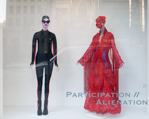 Image of two costumes from the exhibition on mannequins. On the left, a mannequin wears a black wetsuit with red stripes along side the body. It also wears goggles and a swim cap. Next to it, on the right, another mannequin wears an ornate red dress with a red bonnet-like hat.  