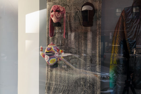 Close-up images of three masks in the exhibition. The first mask on the upper left, is a red and pink crocheted hat with long bunny-like ears and hearts crocheted around the head. There are only two holes for eyes. On the upper left, a mannequin head dons a black ski mask. On the bottom right, a mannequin head wears another crocheted hat and mask shaped like a teapot. The teapot mask is purple, white, pink, red, yellow, and in various hues and includes  one large slit for the eyes. 