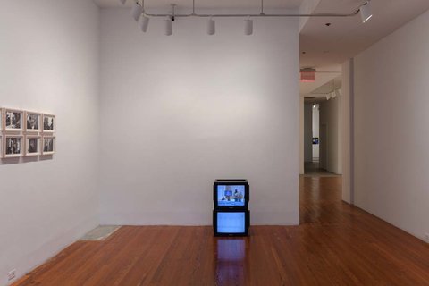 Installation view of the exhibition featuring two walls. On the left wall, several black and white photos are hung in two rows, one on top of the other. On the back wall, in front of it, two monitors, stacked on top of each other. 