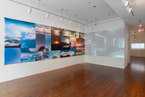 Installation view of two walls in the exhibition. On the left side wall, a collage of several colorful natural landscapes are arranged in three rows. At the corner, the left wall meets the right to blend into a projection of what looks like random lines and shapes. 