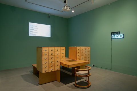 Installation image featuring a gallery room in the exhibition. The walls in this room are painted a forest green. On the left, back wall, a projection is projecting various statistics and numbers about climate change, though they are illegible from the camera point of view. On the right wall, a neon sign is lit with the word 'CLOSED'. Above the word 'CLOSED' is the word, 'OPEN' but it is not lit up. In the middle of the room, there is a group of wooden furniture included a wooden chair, desk, and two large index cabinets with many drawers. The chair faces the project. On the table, in front of the chair, is a tray. 