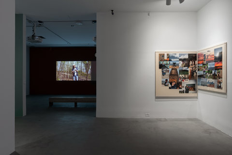 Installation view of the exhibition featuring the right side of a room with a collage of photographs on a white wall. Leading into the back of the image, is a darker room featuring a projection on a dark colored wall. In front of the projection is a wooden bench. 