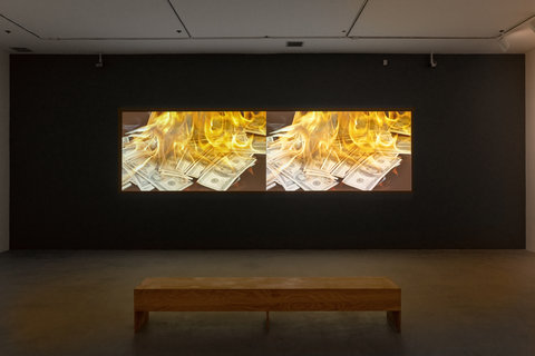 Installation view of two side by side projects against a dark colored wall. The projects, side by side, are identical and feature a pile of money on fire. In front of the projection is a wooden bench. 