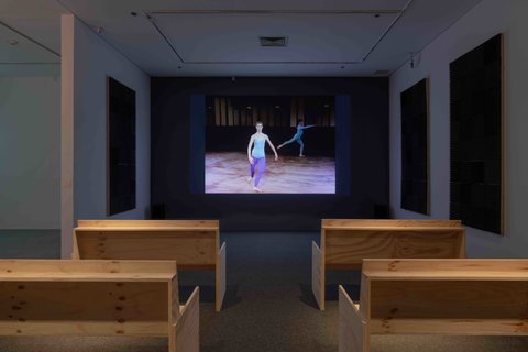 View of the installation of a video in the exhibition. The video is projects against a dark wall at the front of the room. The backs of the wooden benches, arranged like church pews, can be seen from the camera angle at the back of the room. The white walls have large foam pads to absorb the sound. At this moment, the still from the video in the installation view image features a dancer in a blue leotard. In the back of the still, another dancer wears a blue leotard in the shadows. 