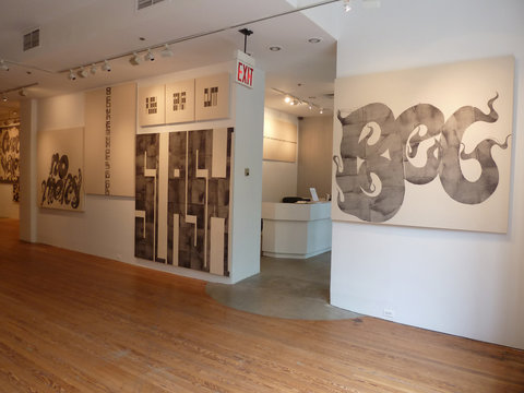 Gallery installation view of the exhibition featuring two walls separated by a doorway that looks into the office space and entrance lobby. On the right is a conglomerate of about ten works of art. The wall on the left has a majority of the artwork. On the left most side, there is a singular drawing. Next to the drawing, on the right side on the top is three framed smaller works. Below those smaller works is a collection of dark drawings. On the right side of the entrance to the gallery, is a large ball point pent drawing of a figure that seems to resemble a snake or a dragon as it's tail and limbs swirl around the page. 