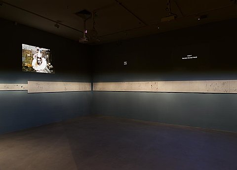 A film is projected on the top half of a darkened gallery wall. A strip of drawings, lighted with direct light, runs along the middle of the wall.