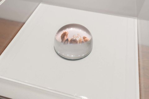 Close-up image of a glass orb in the exhibition atop a white pedestal. The glass orb's convex shape allows for the image underneath to show through in a bit of a distorted way. 