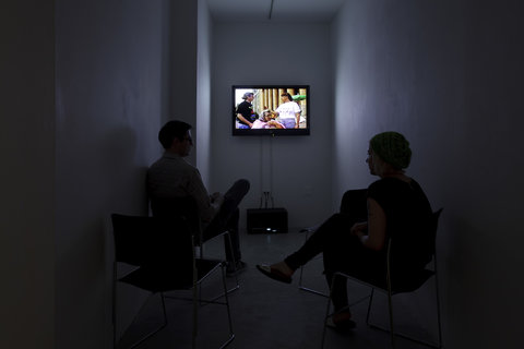 Two people are seated in a dark room facing a television screen that is mounted on the wall. On the screen are three figures. The television is the only source of light in the room.