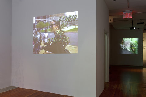 A projection on a white wall. The projection features a woman and the words "Studio of the Streets" in yellow writing over the image. In the background, a darker room features a different projection. In this projection a woman in a green winter coat faces a figure whose back is to the camera.