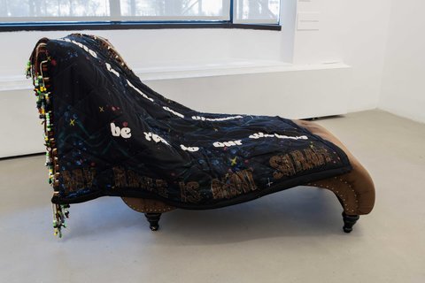 Installation view of the sculpture in the exhibition in the middle of the floor. It appears to be a dark brown lounger chair covered by a large black piece of textile with beads and the text “be real to our women”, and “MY AUNT IS AUNT SARAH”.