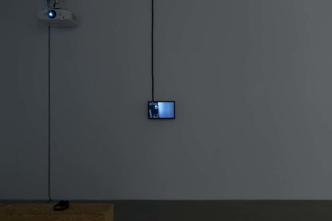 Installation view of a small television monitor mounted to a white wall. The lights are off in the room and the television monitor gives off a bluish glow from the still on the screen from the video. 