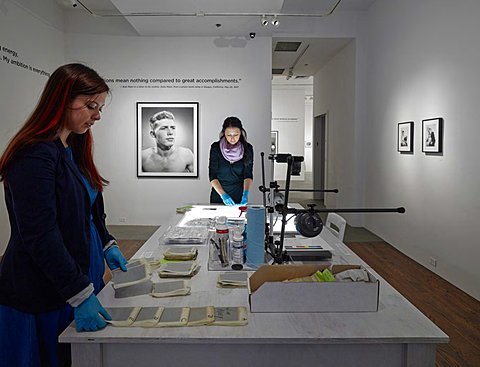 There is a room with a table in its center. The table is filled with unprinted photographs and negatives that two students are working to develop. On the right parallel wall to the table there are two small framed black and white photographs side by side. The back wall perpendicular to the table has a larger framed black and white portrait photograph of a man. Above the photograph of the man is a quote that wraps around onto the left parallel wall to the table.