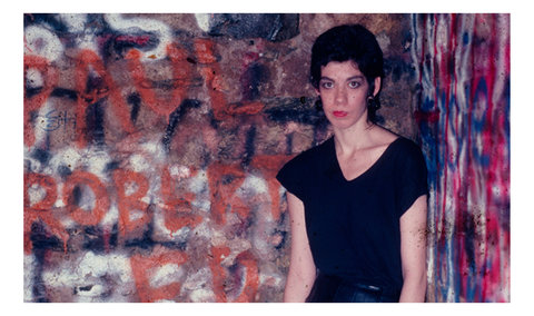 Image of a figure in a black shirt, with short cropped black hair, and red lips, leaning against a wall of graffiti paint.