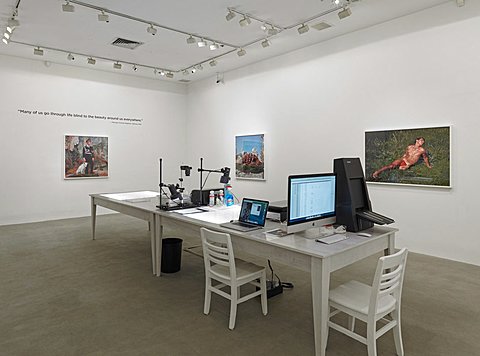 There is a room with two tables spanning the center of the floor. The table is white and there are two chairs in front of two computers that are placed on top of the table. Also on the table is equipment for developing and printing photographs. On the right parallel wall to the table there are two framed photographs. The one to the right depicts a man laying in foliage while the left one depicts a landscape, both are in color. On the perpendicular wall to the table there is one colorful portrait photograph centered on the wall, this is also in color. Above this picture is a quote written in black text. 