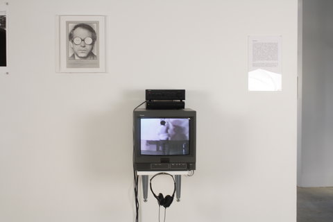 Gallery installation view of a TV in the middle of the wall. On the television is a black and white still. A pair of headphones hangs from the front of the TV. On the top left corner, hanging above the television is a black and white picture of a man with short hair wearing eyeglasses that cover his eyes. He is straight-faced with no emotion. Wall text to accompany the exhibition is on the right side. It is illegible from the image. 
