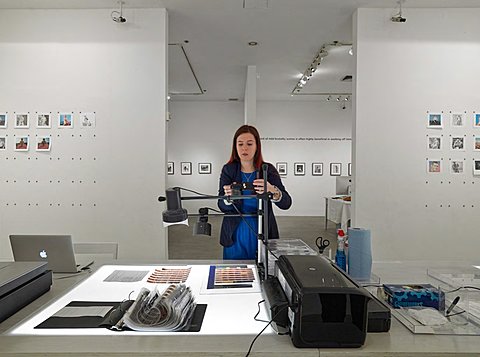 A student examines photographic film sheets on top a Lightbox. On the wall behind her there are small square photographs tacked to the wall.