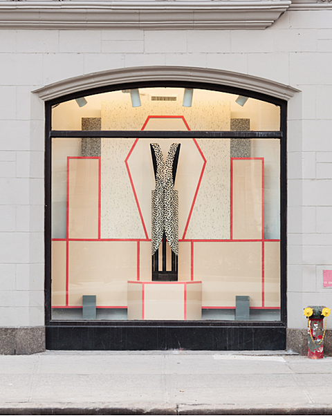 A cheetah print smock like textile is hanging and draped over a small brown table structure. This is placed on top of a tan colored round pedestal that is lined and divided into three visible sections with red lines. Behind the garment are granite like slabs of backdrops and tan boarding geometrically divided by red lines in the same fashion as the pedestal. 