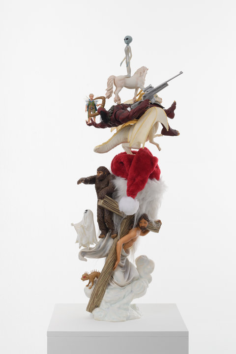 Close-up sculpture in the exhibition featuring a stack of figurines. From top to bottom, the figurines are a small white, shiny alien with a large balloon head and dark teardrop eyes, standing atop a white unicorn, which is standing atop a dark red, satanic figure with fiery eyes and massive horns. The figure is laying flat and its legs are on the shoulders of a white dinosaur-like figure with big yellow horns and big wings. The Satanic figure's palms are outstretched and in its palm holds a small nymph-fairy figure. The dinosaur stands atop a Santa Claus with a classic red hat, long white beard, and wire-rimmed oval glasses. On the back of Santa's head is attached a bigfoot figurine and a small white sheet ghost figurine. At the base of these figurines a Jesus holding a big wooden cross in a white toga. On the bottom of the toga, is a small red-eyed reptilian creature figurine. The very bottom base appears to be white, cloud-like shapes. 