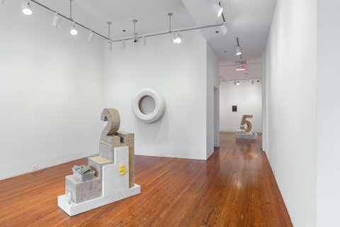 Gallery view of the exhibition featuring gallery one, and the hallway that leads into gallery two. On the ground, in the middle of the gallery before the hallway, is a tile sculpture of the number two. Behind the sculpture, is a white fiberglass tire. On the tire are light orange ginko leaf stamps. Leading into the hallway, into gallery two in the distance, there is a tile sculpture of the number 5. 