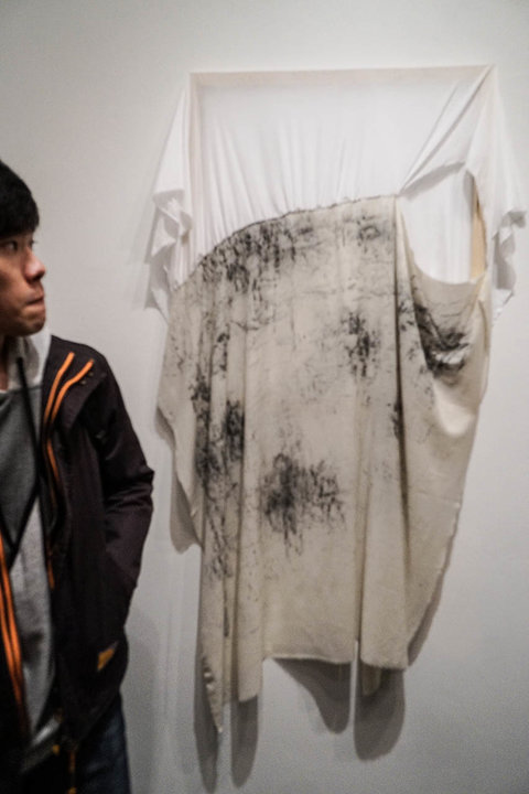 Installation image of a draped fabric hanging on the wall in the exhibition. The white sheet, has black stains speckled about. 