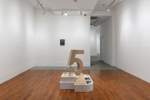 Gallery view of the exhibition. In the middle, on the floor of the gallery is a tile sculpture of the number five. Behind it on the wall is a small drawing and next to the drawing is the hallway leading into gallery three.  