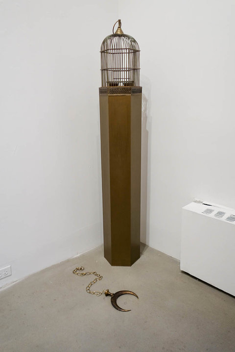 Installation image of a sculpture in the corner room of the exhibition. The sculpture features a bronze-colored podium or pedestal. On top of the pedestal sits a bronze-colored bird cage. On the ground in front of the pedestal, a chain with a crescent moon in the same bronze hue sits on the floor.  