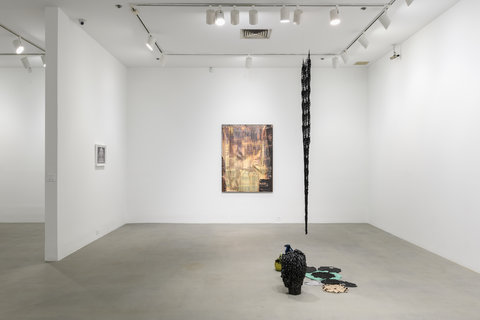 Gallery installation image featuring a profile view of a hanging metal crocheted mesh sculpture. It is think and looks like a line from the side view. Below it sits a dark colored ceramic vase. In the background is a multi-colored, beige and purple work on the wall. It is shiny and reflects the gallery light. On the left wall is a small drawing that is illegible from the viewpoint of the camera. 