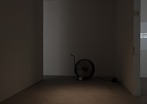 A black fan on wheels with a handle in a dark corner of the gallery.